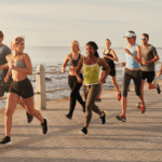 Grupo de corredores de rua em movimento durante uma prova ao ar livre, vestindo roupas esportivas leves e coloridas. A imagem mostra atletas de diferentes perfis correndo juntos, com foco na energia, determinação e espírito coletivo, em um cenário de praia ao fundo.