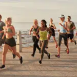 Grupo de corredores de rua em movimento durante uma prova ao ar livre, vestindo roupas esportivas leves e coloridas. A imagem mostra atletas de diferentes perfis correndo juntos, com foco na energia, determinação e espírito coletivo, em um cenário de praia ao fundo.