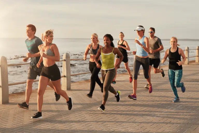 Grupo de corredores de rua em movimento durante uma prova ao ar livre, vestindo roupas esportivas leves e coloridas. A imagem mostra atletas de diferentes perfis correndo juntos, com foco na energia, determinação e espírito coletivo, em um cenário de praia ao fundo.