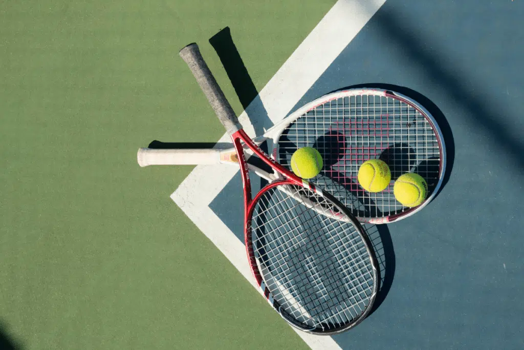 Duas raquetes de tênis apoiadas no chão da quadra, com bolas de tênis espalhadas ao redor, sobre o piso com marcações brancas visíveis. A cena transmite preparação para treino ou partida, com iluminação natural destacando a textura da quadra e o clima esportivo do ambiente.
