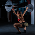 Mulher realizando levantamento de peso olímpico (LPO) na academia, executando movimento com barra e anilhas acima da cabeça, demonstrando força, técnica e potência em treino de alta performance.