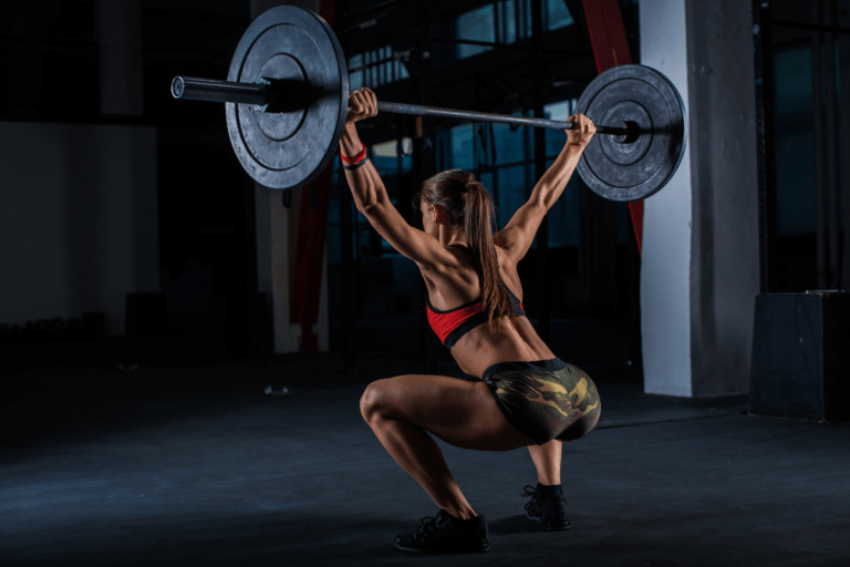 Mulher realizando levantamento de peso olímpico (LPO) na academia, executando movimento com barra e anilhas acima da cabeça, demonstrando força, técnica e potência em treino de alta performance.