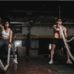 Duas pessoas realizando exercício com corda naval em um box de treinamento funcional, executando movimentos intensos e sincronizados, demonstrando força, resistência e alto gasto calórico em treino de alta intensidade.