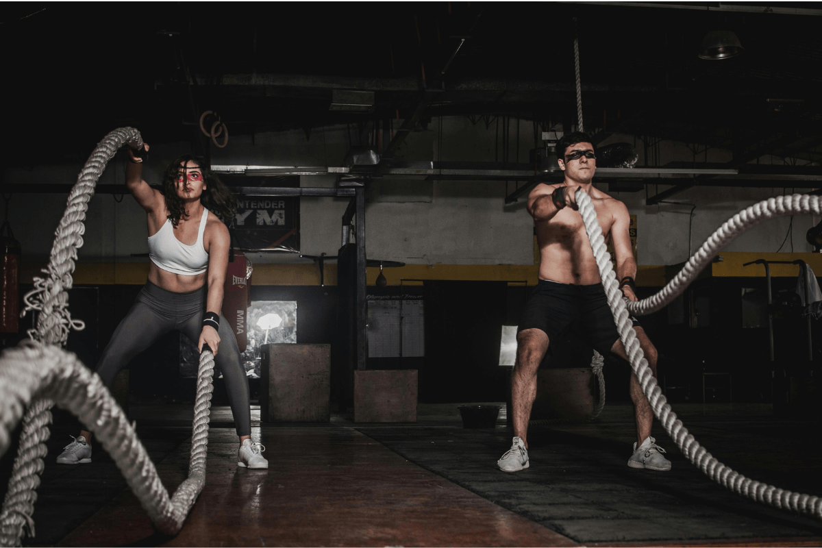 Duas pessoas realizando exercício com corda naval em um box de treinamento funcional, executando movimentos intensos e sincronizados, demonstrando força, resistência e alto gasto calórico em treino de alta intensidade.