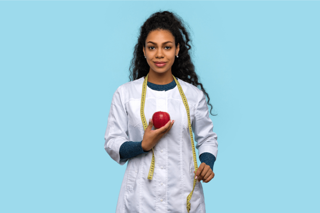 Nutricionista mulher em pé, usando jaleco branco, segurando uma maçã vermelha e uma fita métrica nas mãos, transmitindo saúde, profissionalismo e foco em nutrição e qualidade de vida.