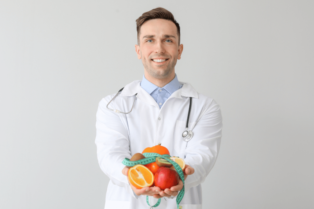 Nutricionista homem em pé, usando jaleco branco, segurando frutas e uma fita métrica nas mãos, transmitindo saúde, profissionalismo e foco em nutrição e qualidade de vida.